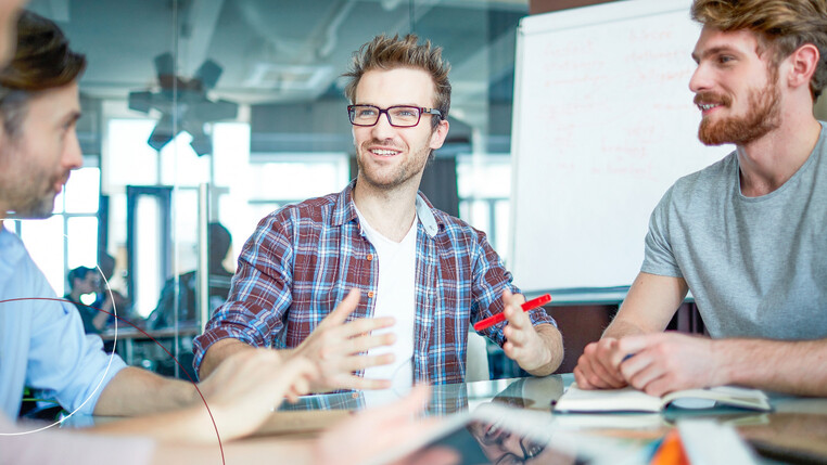 Drei Männer sitzen an einem Tisch und unterhalten sich, im Hintergrund eine Flipchart