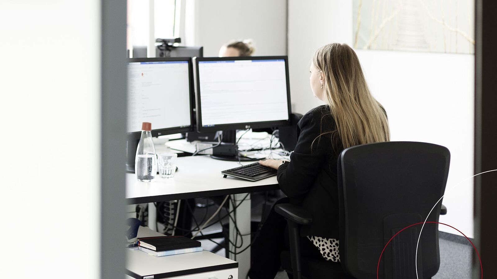 Blick von hinten auf eine vor zwei Monitoren sitzende Mitarbeiterin