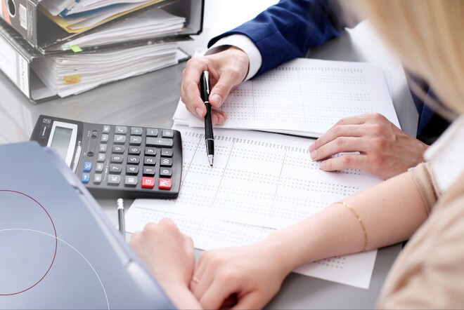 Eine Frau und ein Mann sitzen an einem Schreibtisch und beugen sich über einen Taschenrechner und ihre Zahlen.
