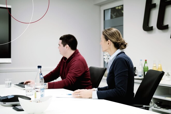 Beate Theede und Mitarbeiter sitzen an Konferenztisch und blicken an Wand