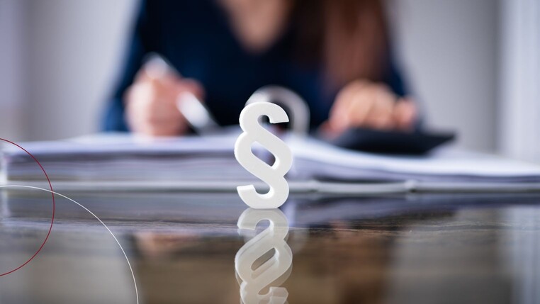 Stehender Paragraph vor einer arbeitenden Frau am Schreibtisch.