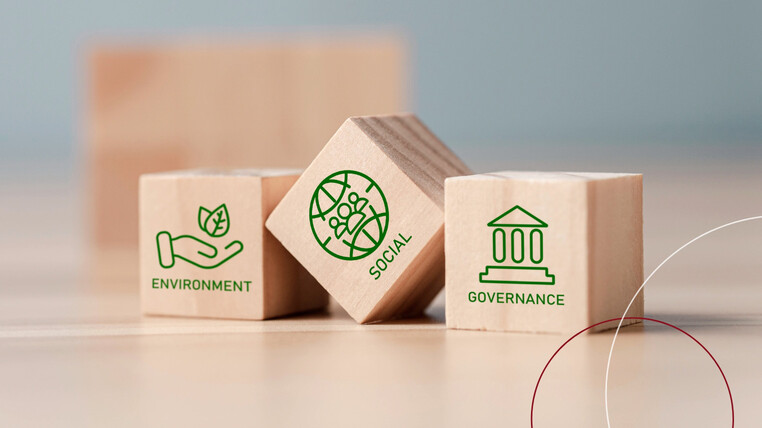 Drei Holzwürfel liegen auf einem Tisch. Jeder Würfel hat ein Wort und ein Bild auf der Frontseite. Würfel 1: Environment mit einer Hand und Blättern darüber; Würfel 2: eine Weltkugel mit Menscehn, darunter das Wort Social; Würfel 3: ein Säulengebäude grafisch dargestellt, darunter das Wort Governance.