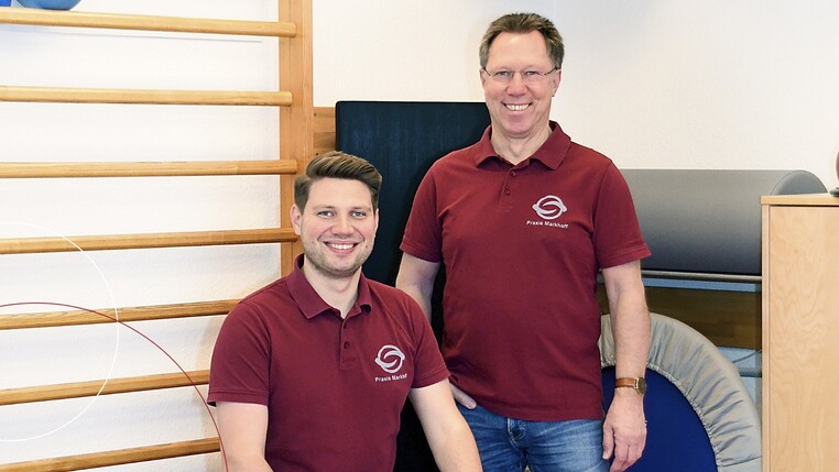 Jens-Peter Markhoff und sein Sohn Lasse stehen bzw. sitzen vor Trainingsgeräten, wie der Sprossenwand und einem Trampolin und lächeln in die Kamera.