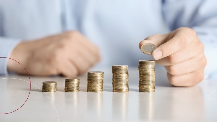 Unterschiedlich hoch gestapelte Geldmünzen stehen auf einem Tisch vor einer Person.